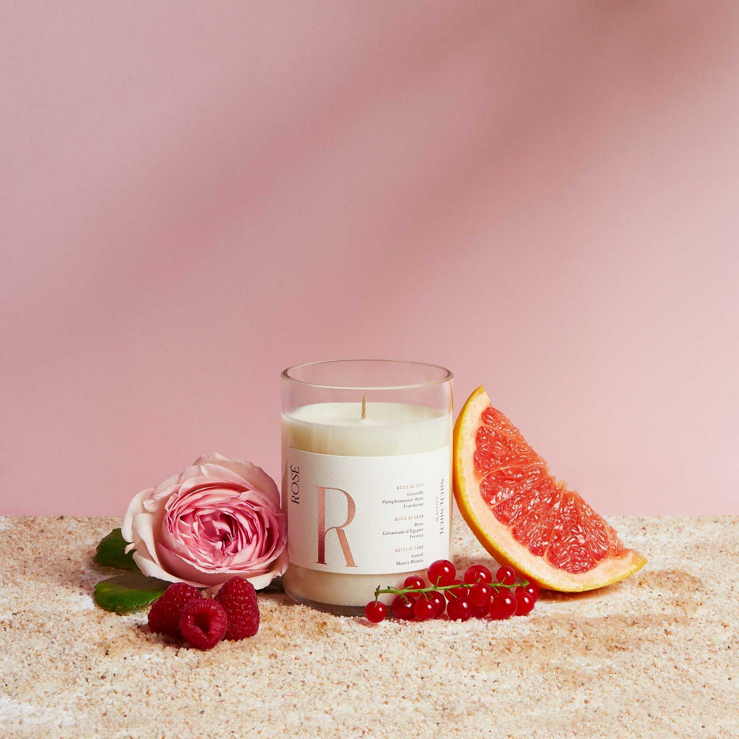 Maison Tchin Tchin bougie parfumée senteur rosé posée sur du sable à côté des fruits rouges et d'une rose qui composent son parfum