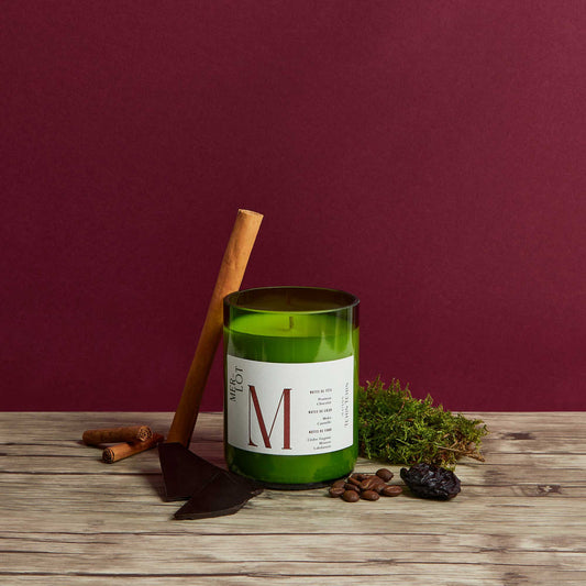 Bougie naturelle à la cire végétale au parfum inspiré par le merlot disposé sur une planche à côté de bâtons de cannelle, de morceaux de chocolat et de grains de café