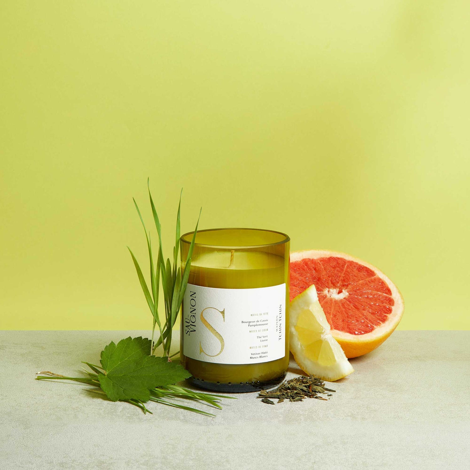 Maison Tchin Tchin bougie artisanale parfumée au vin blanc fabriquée en France aux arômes du sauvignon posée à côté d'agrumes et d'herbes