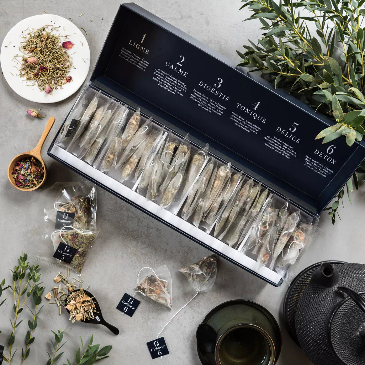 Coffret cadeaux de 36 sachets d'infusions ouvert avec des plantes et une théière posés sur une table.