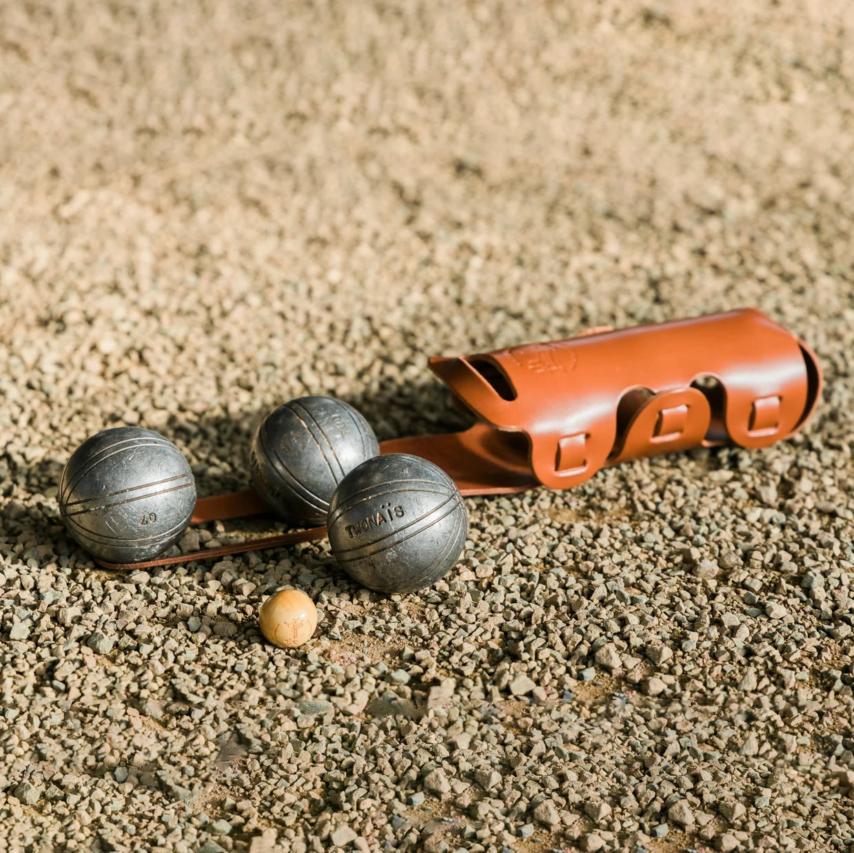 Etui à pétanque en cuir posé sur le sol avec les boules et le cochonnet à côté